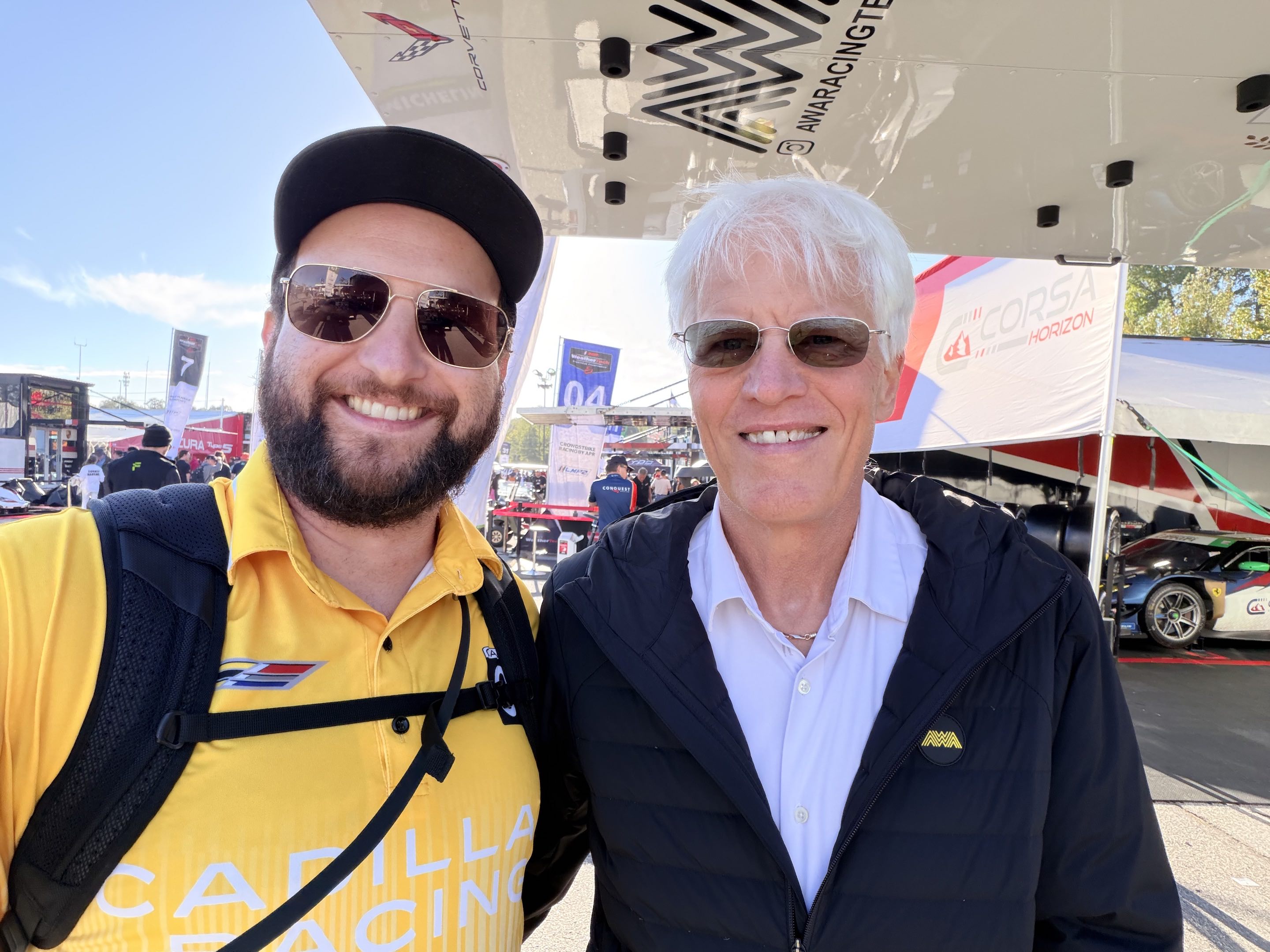An old man and an older man, both wearing sunglasses, posing in front of a garage in a racing paddock